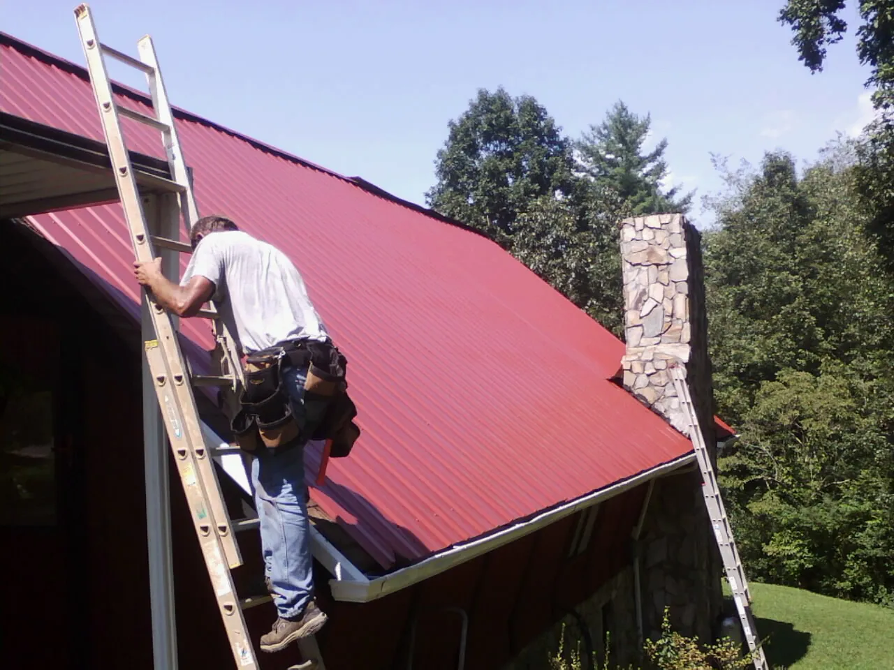 Roofer on a ladder beside a red metal roof and stone chimney — Soffit & Fascia Repair by Roofing Brothers in Hot Springs